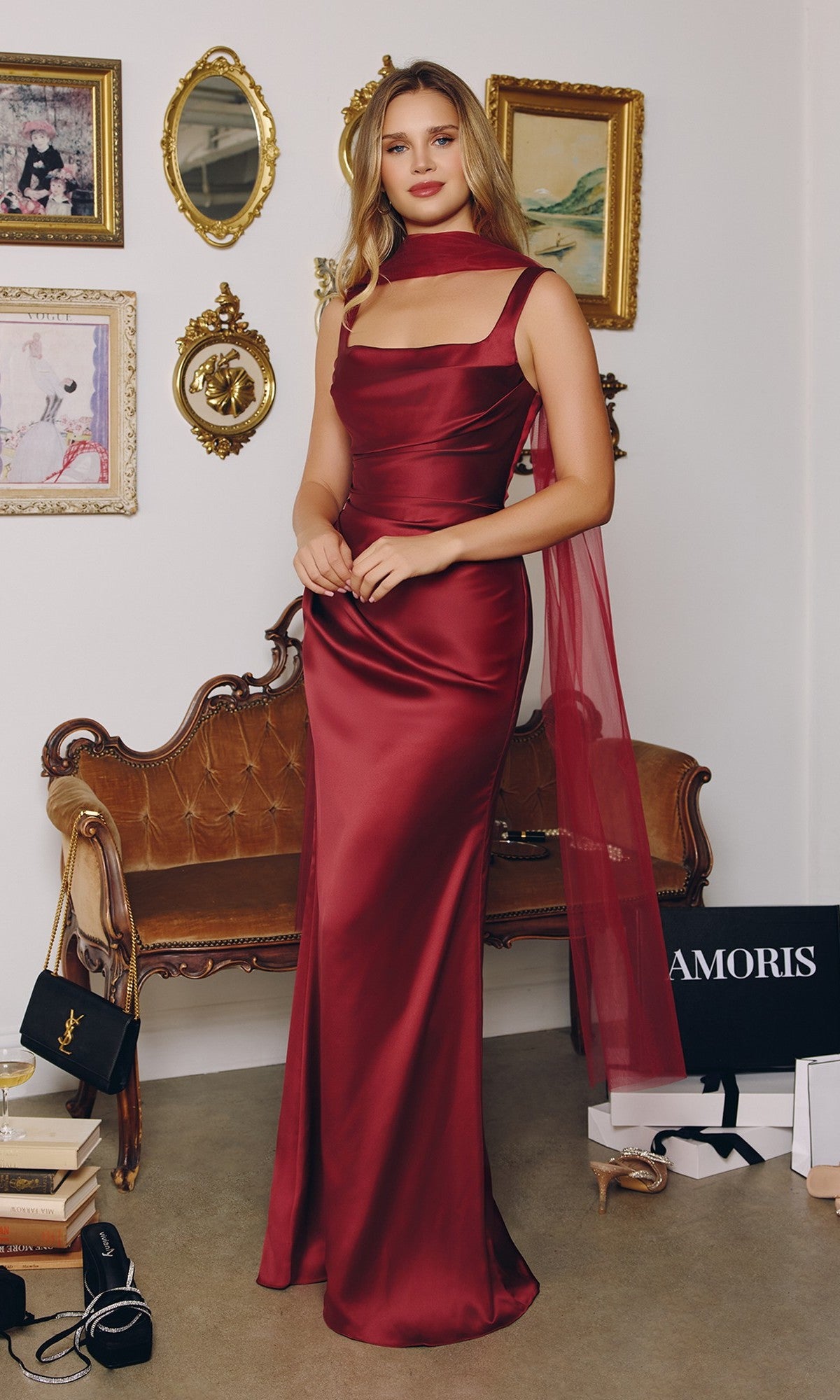 A woman wearing the Amoris T729 long burgundy prom dress with a square neckline stands in a decorated room featuring framed art, a vintage sofa, and shopping bags on the floor.
